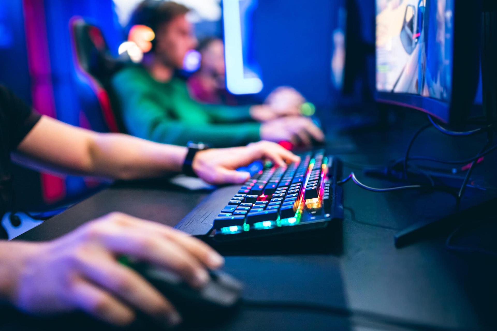 Hands on a glowing RGB keyboard and mouse, with gamers in headsets blurred in the background of a neon-lit room