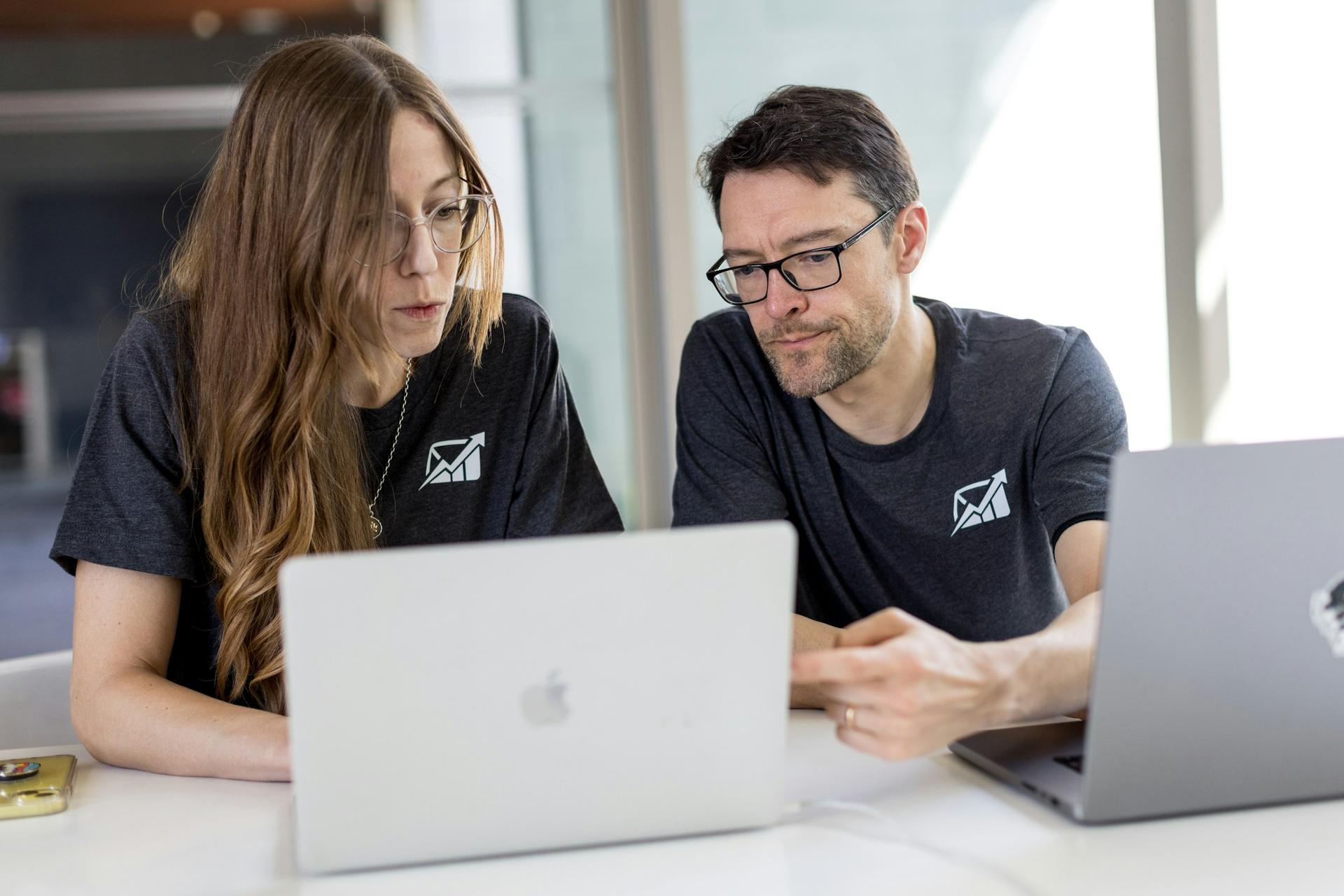 Two coworkers in matching T-shirts leaning in to review something together on a laptop at a bright office table