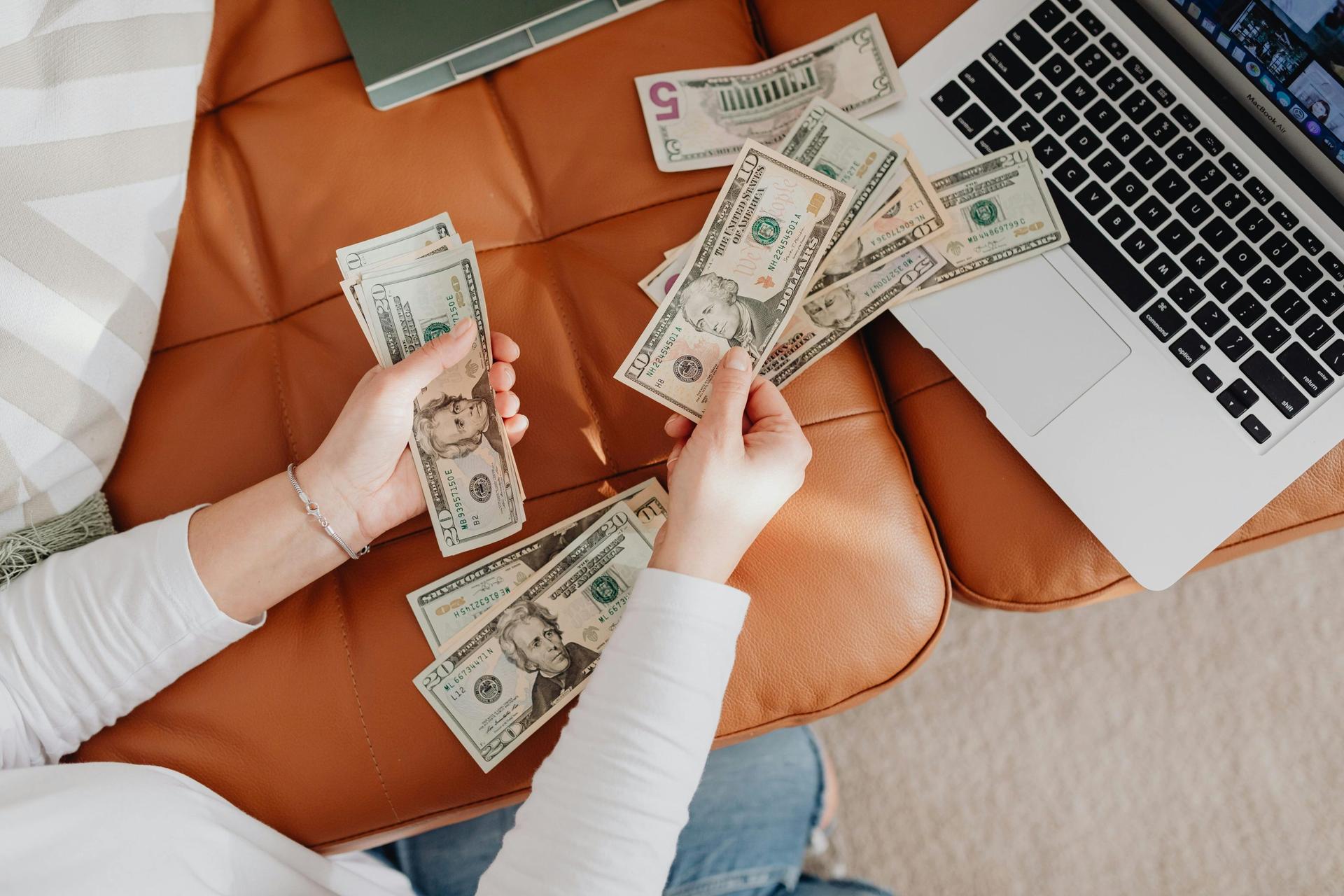 Overhead view of hands counting $20 and $10 bills on a leather chair next to an open MacBook Air